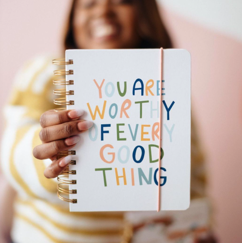 black woman in the background and she is holding a white notebook in the foreground that says, "You are worthy of every good thing"