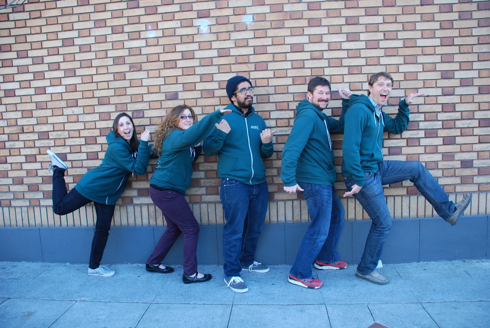 5 people in green hoodies posing silly against a brick wall